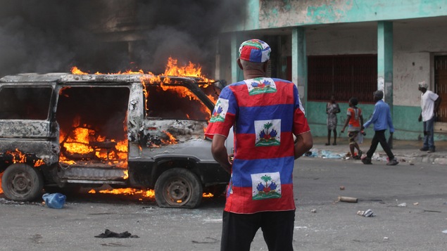 Un homme vêtu d'une tunique aux couleurs nationales se tient devant une fourgonnette en flammes dans une rue haïtienne.