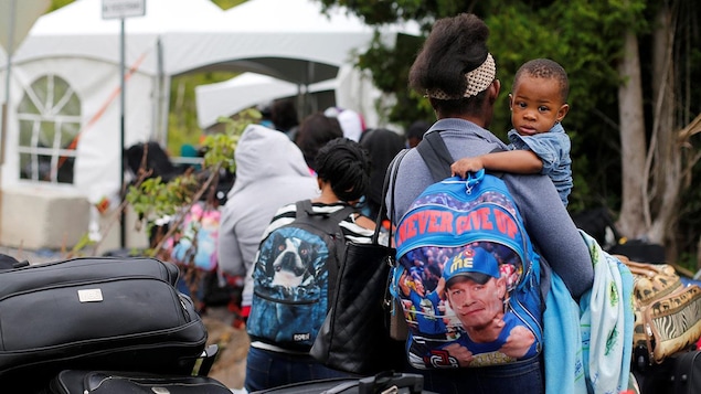 Un petit garçon aux bras de sa maman qui soutient un sac à dos "Never give up" dans une file