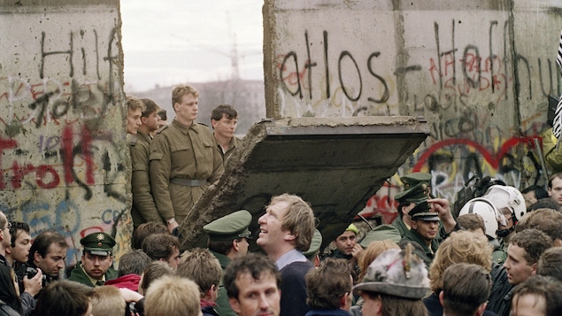 Des gens du côté ouest se massent le long du mur en novembre 1989, sous l’œil des soldats de Berlin-Est, alors qu'un pan du mur s'effondre.