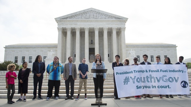 Quatorze jeunes dont une qui parle au microphone et sept qui tiennent une grande affiche, devant les marches de la Cour Suprême des États-Unis à Washington.