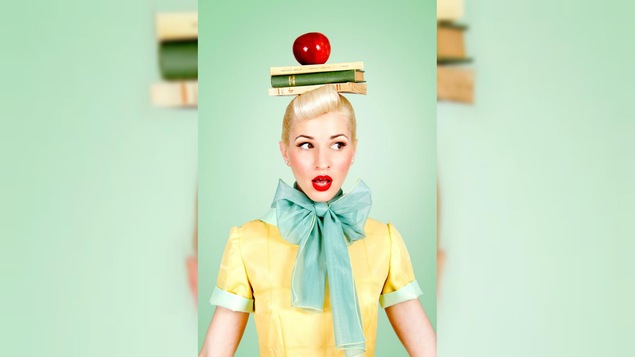 Une femme costumée dans le style pin up tient des livres et une pomme en équilibre sur sa tête.