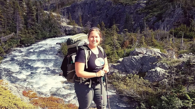 Une femme en randonnée pose pour la caméra sur un mont, près d'un cours d'eau.