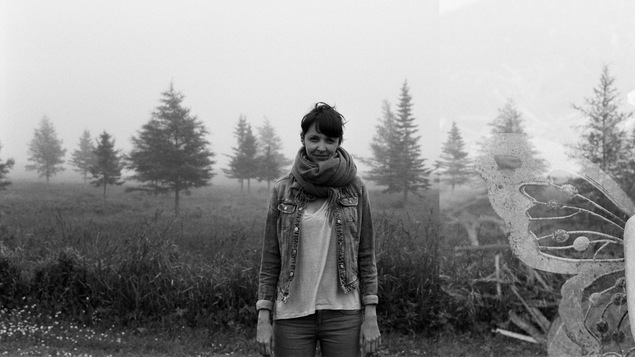 Portrait en noir et blanc de l'auteure Kristina G. Landry, debout, en extérieur, devant une rangée d'arbres, les bras le long du corps. Elle porte un t-shirt clair, une veste en jean et un gros foulard autour du cou.