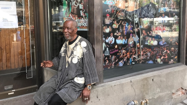 Un homme fume la cigarette alors qu'il est assis sur le rebord de la fenêtre d'un local commercial. Dans la vitrine sont présentées des photos de musiciens africains.