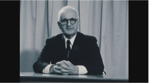 Image en noir et blanc d'un homme portant des lunettes assis.