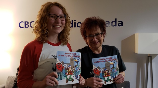 Deux femmes tiennent chacune dans leurs mains un exemplaire en anglais et en français du livre jeunesse Monica-Claire au Festival du Voyageur.