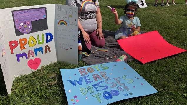 Une femme et un enfant, portant masques et lunettes fumées, sont sur la pelouse devant deux grandes affiches qui disent « Fière maman » et « Les garçons portent aussi des robes ».