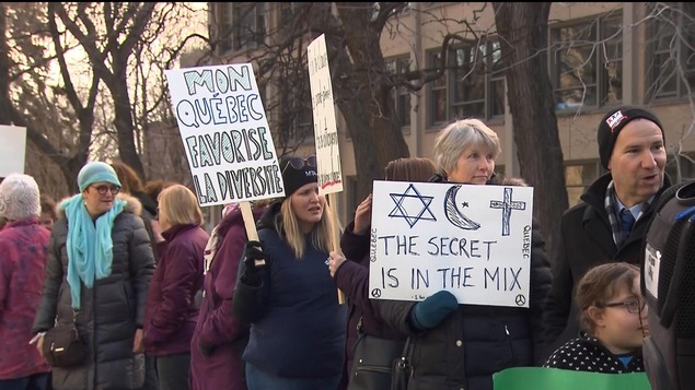 Des personnes portent des pancartes sur lesquelles est écrit : « Mon Québec favorise la diversité » et «The secret is in the mix », cette dernière accompagnée de dessins représentant une croix de David, un croissant et une étoile, ainsi qu'un crucifix.