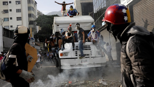 Des manifestants portant des masques à gaz et des casques sont montés sur un camion. Ils sont entourés d'une fumée blanche, qui est en fait du gaz.