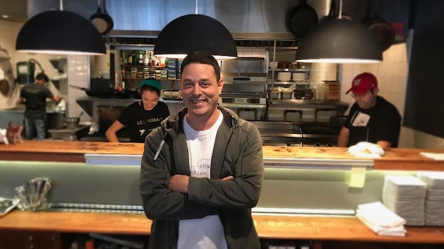 Le restaurateur est au centre de l'image. À l'arrière-plan, des cuisiniers s'affairent devant des appareils de cuisine.