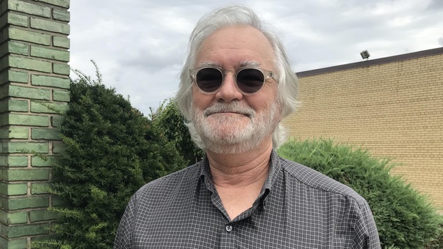 Photo d'un homme souriant aux cheveux et barbe blancs qui porte des lunettes fumées