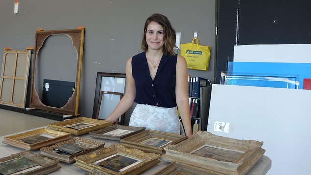 La photo montre une jeune femme dans un atelier, prenant la pose devant une table sur laquelle sont posés des oeuvres encadrées.