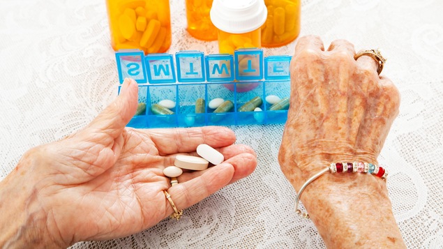 Les mains d'une vieille dames tiennent des médicaments variés. En arrière-plan, on voit un pilulier et des contenants de médicaments.