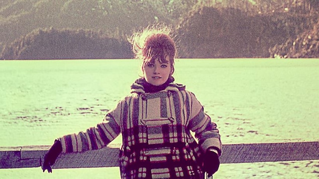Une femme en manteau pose devant des montagnes et un lac.