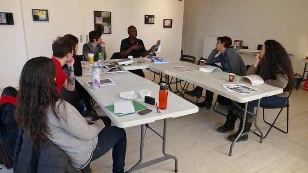 Six personnes assises dans un atelier écoutent Wilgis Agossa décrire une photo. Six personnes assises dans un atelier écoutent Wilgis Agossa décrire une photo.