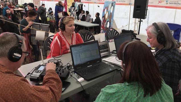 Assise devant une table, Paulette porte un chandail rouge et blanc, elle fait face à l'équipe de l'émission: Animatrice, technicien et journaliste.