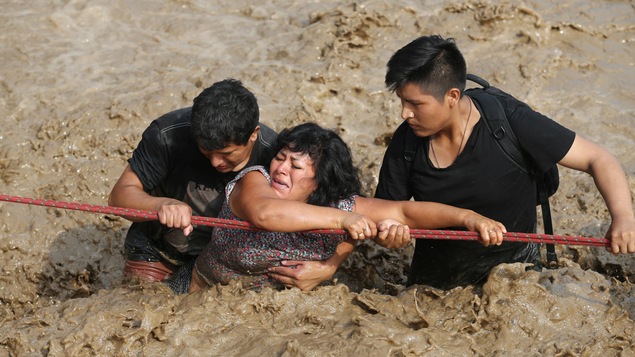 Deux personnes aident une femme à traverser une rue inondée après le débordement d'une rivière à Huachipa, au Pérou. Deux personnes aident une femme à traverser une rue inondée après le débordement d'une rivière à Huachipa, au Pérou.