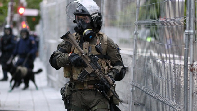 Un policier transportant une arme d’assaut dans ses mains monte la garde devant une clôture grillagée. Il porte un uniforme de couleur kaki, un casque de protection noir muni d’une visière, un masque à gaz, des jambières et des protège-coudes. À l’arrière-plan, on aperçoit deux autres policiers. L’un d’eux tient un chien en laisse.