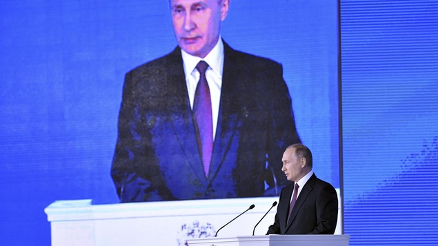 Vladimir Poutine devant un lutrin et un écran géant projetant sa silhouette.