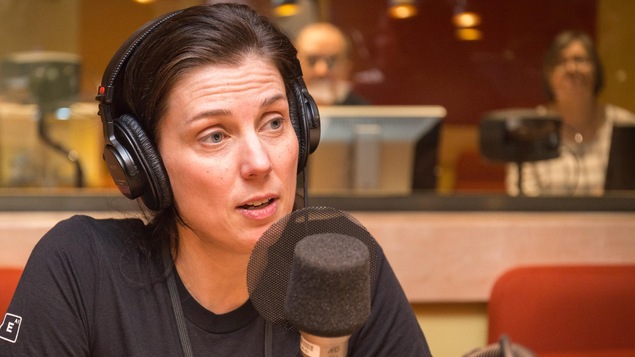 Une photo de Valérie Bécaert, vêtue d'un t-shirt noir et portant des écouteurs recouvrant ses oreilles en train de parler à un micro dans le studio de l'émission La sphère.