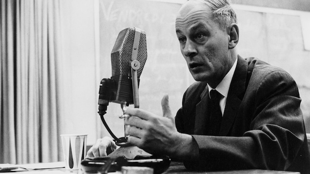 Dans une salle, René Lévesque, ministre des Richesses naturelles et ministre de la Famille et du Bien-être social, est assis à un bureau, une cigarette à la main, derrière un micro sur pied.