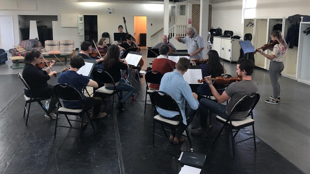 Les membres d'un orchestre sont assis sur des chaises dans un sous-sol devant un chef d'orchestre.