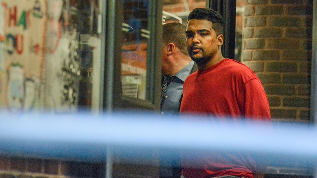 Un jeune homme, qui semble blessé à la tête, porte un chandail rouge et est escorté par un policier. Un jeune homme, qui semble blessé à la tête, porte un chandail rouge et est escorté par un policier.