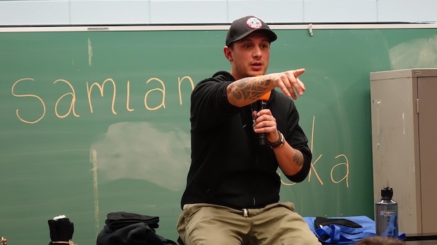 Le rappeur, poète, acteur, photographe et conférencier Samian, assis sur un bureau, micro à la main, lors d'une rencontre avec les jeunes de l'école secondaire Otapi, à Manawan en février 2018.