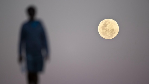 Un rond lumineux, la Lune, est photographié dans le ciel. La silhouette d'une personne est vue à gauche de l'astre.
