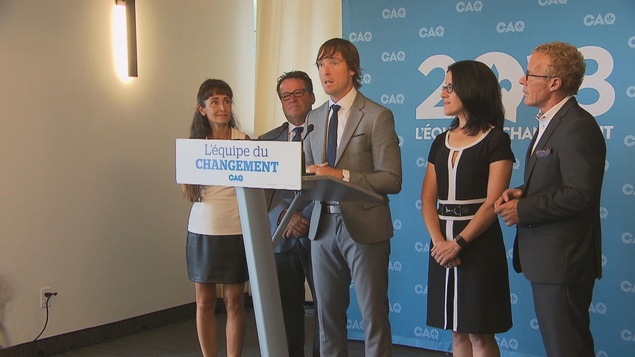 Trois hommes et deux femmes, debout derrière un lutrin. L'homme au centre parle.