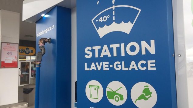 Une pompe, bleue, avec des pictogrammes et les inscriptions : « Station lave-glace », « payez », « ouvrez » et « remplissez ».