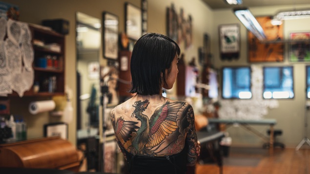Une femme de dos dans un salon de tatouage exhibe un dragon coloré dessiné sur son dos.
