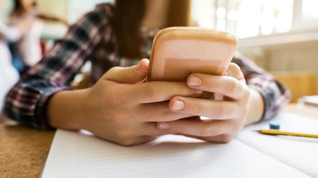 Un gros plan sur les mains d'une jeune fille qui tient un téléphone intelligent. Ses mains sont posées sur son pupitre d'école, par dessus un cahier ligné.