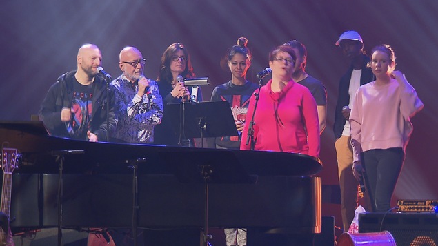 On voit des artistes avec un micro en main. Il sont positionnés derrière un piano à queue sur une scène d'une salle de spectacle.
