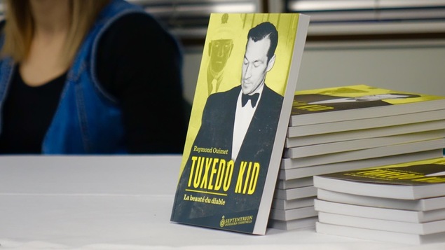 Une pile de livre empilés sur une table. Un des livres est accoté sur les autres. Sur la couverture, la photo en noir et blanc d'un homme en costume et en nœud papillon. On peut y lire « Tuxedo Kid, la beauté du diable. »