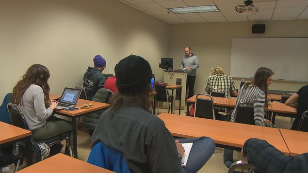 Étudiants de dos dans une salle de classe devant un professeur.