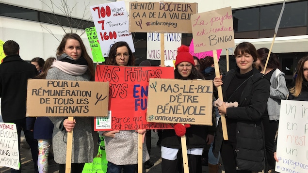 Quatre étudiantes tiennent des pancartes où des slogans sont inscrits. Il peut y lire «Pour la rémunération de tous les internats» et «La psycho est rendue un bingo»