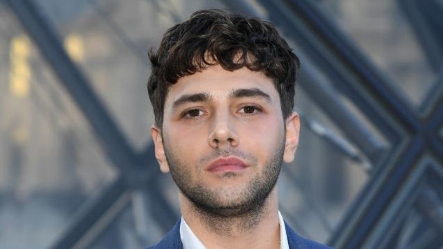 Xavier Dolan porte un costume bleu et une chemise blanche.