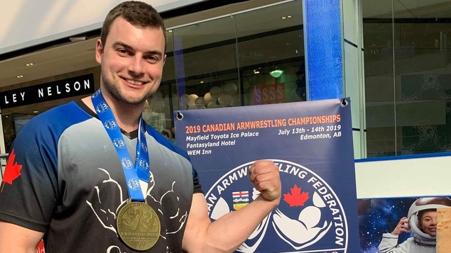 Un homme portant une médaille d'or au cou pose devant l'affiche des championnats du monde de tir au poignet d'Edmonton.