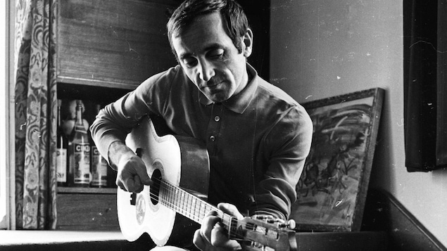 Charles Aznavour joue de la guitare, assis sur des marches, dans une maison.