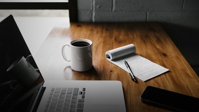 On y voit un ordinateur portable, une tasse de café, un carnet de notes et un cellulaire sur un bureau de travail.