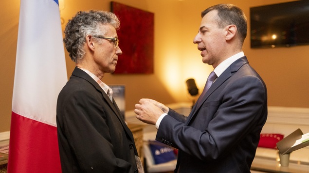 Une homme recoit une medaille d'un autre homme dans un bureau avec un drapeau de la France.