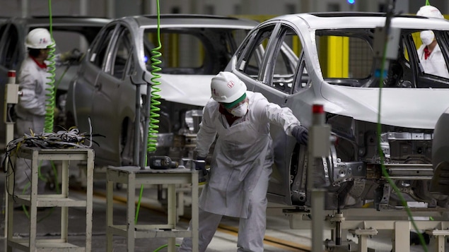 Des employés avec un casque sur la tête et des habits blancs travaillent dans une usine devant trois carrosseries de voiture.