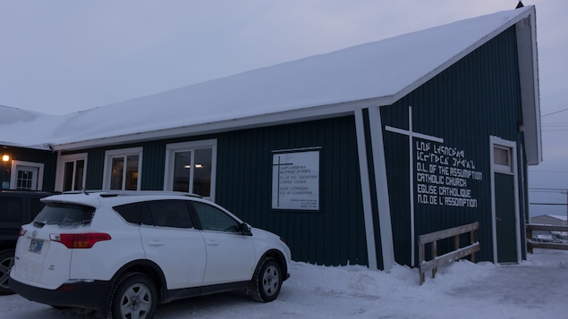 Un bâtiment avec une grande croix sur son toit et une autre sur la façade avec l'inscription « Église catholique Notre-Dame-de-l'Assomption » écrite en inuit, en anglais et en français.