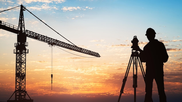 Silhouette Of A Surveyor Standing With Equipment Near Crane At Construction Site During Sunset