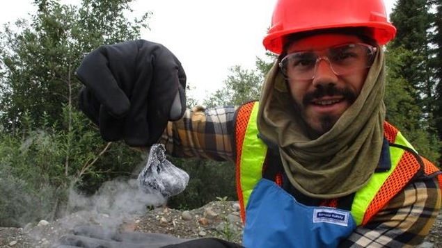 Un chercheur est dans la foret avec un casque route et tient avec des gros gants un objet qui fume.