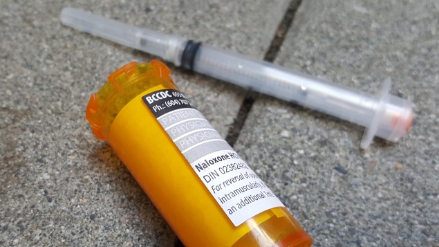 Une seringue usée et un flacon ouvert sur lequel est apposé une ordonnance pour Naloxone sont sur le pavé.