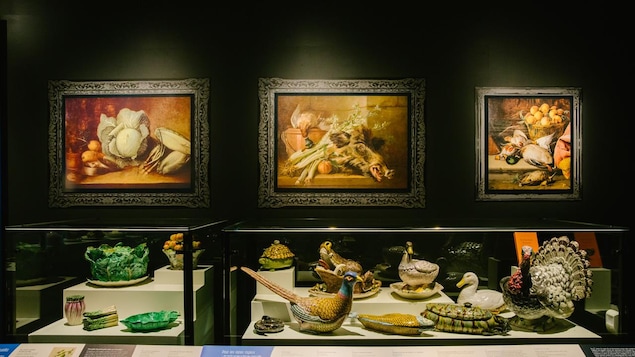 Des plats de service de fantaisie sur un présentoir couvert de verre, surplombé de trois peintures représentant des plats français typiques.