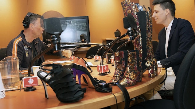 Dans un studio de radio, un animateur parle à un homme. Des chaussures pour femmes se trouvent sur la table.