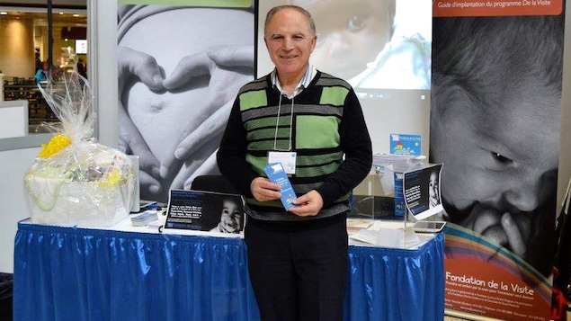 Un homme debout regarde la caméra en souriant devant un kiosque et en tenant une brochure dans ses mains.